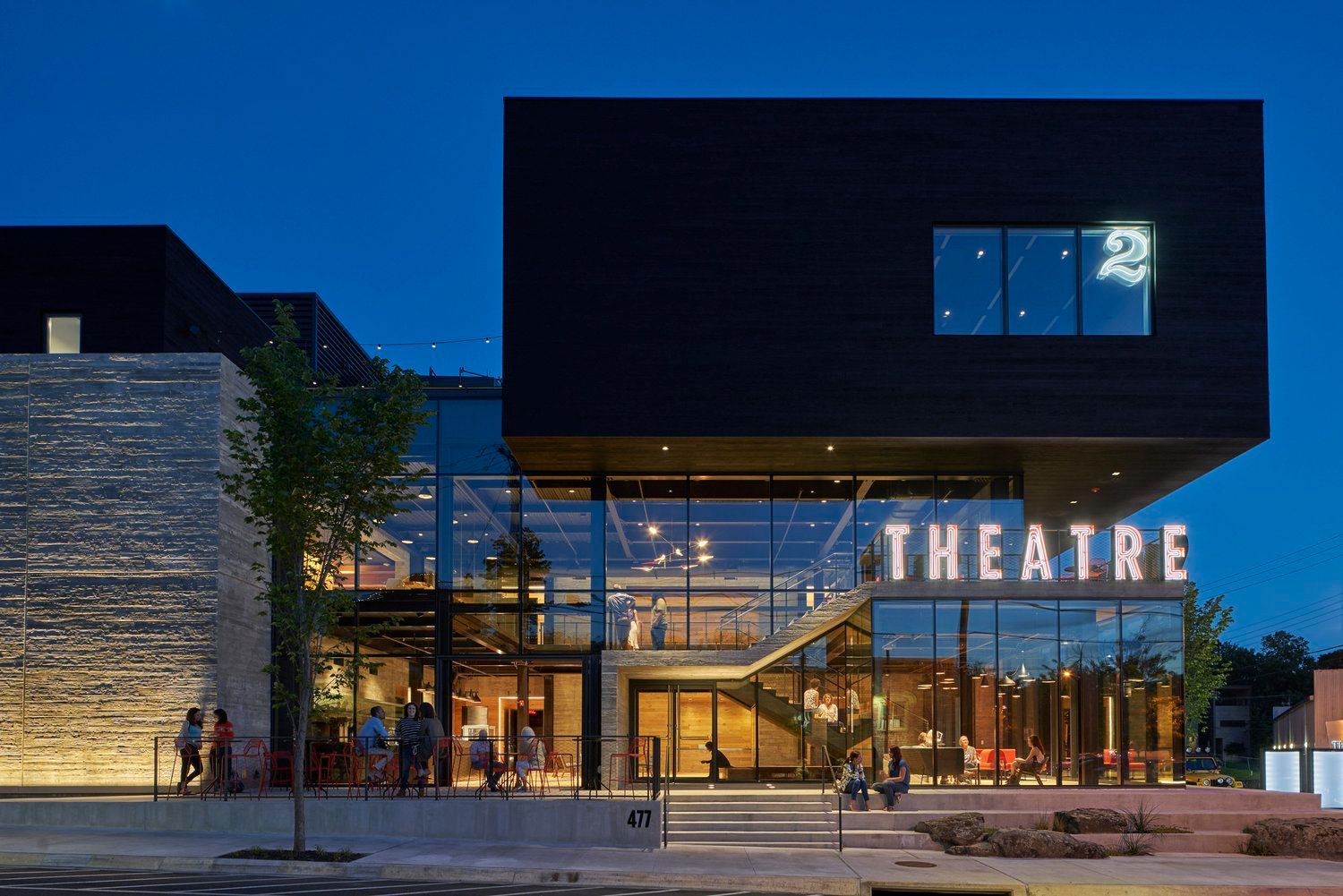 Exterior of a modern theatre building at night, featuring a dark upper section, glass-walled lobby, and stone wall.