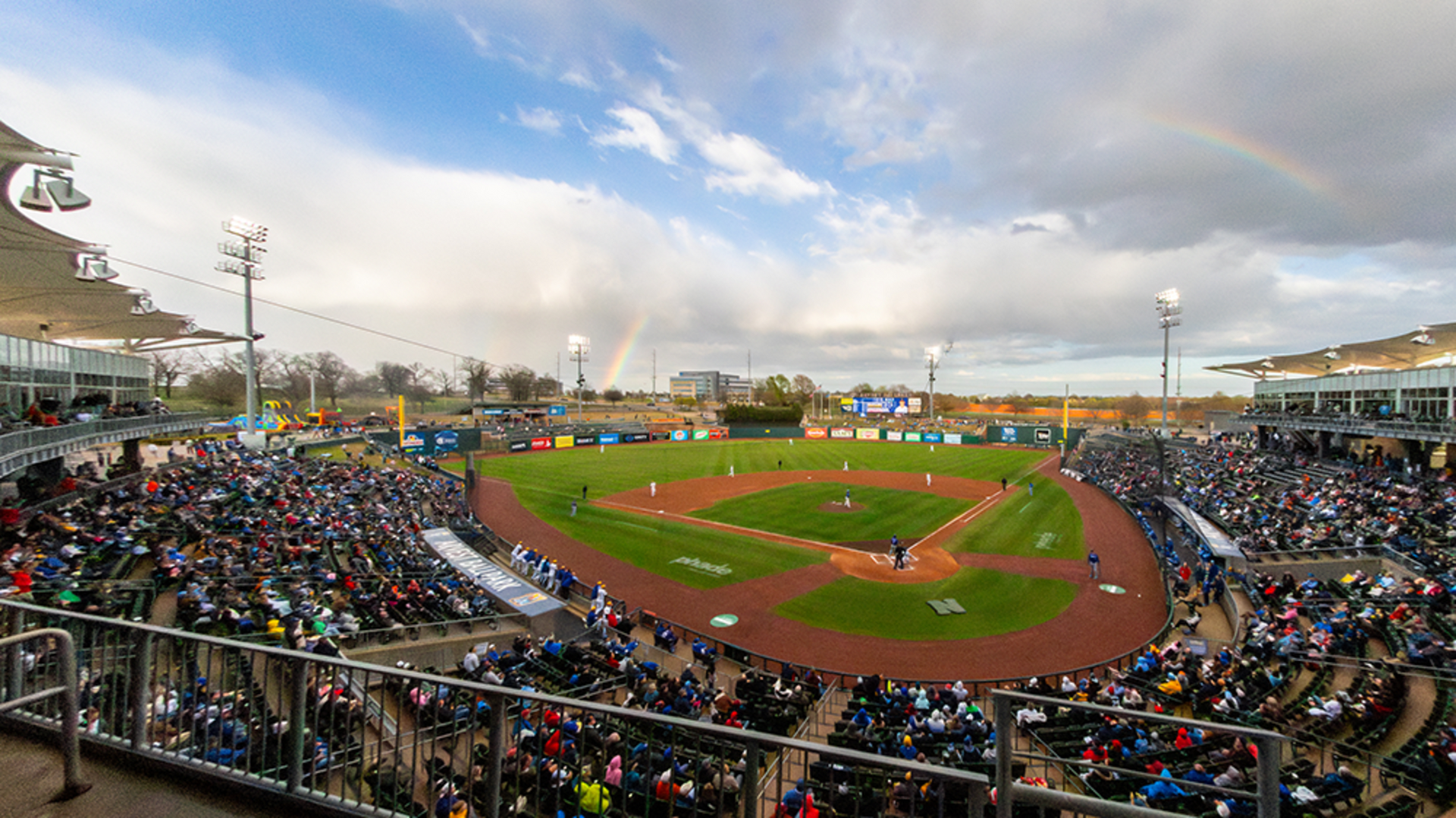 A wide-angle view of a baseball game in a stadium under a cloudy sky with a faint rainbow visible in the distance.