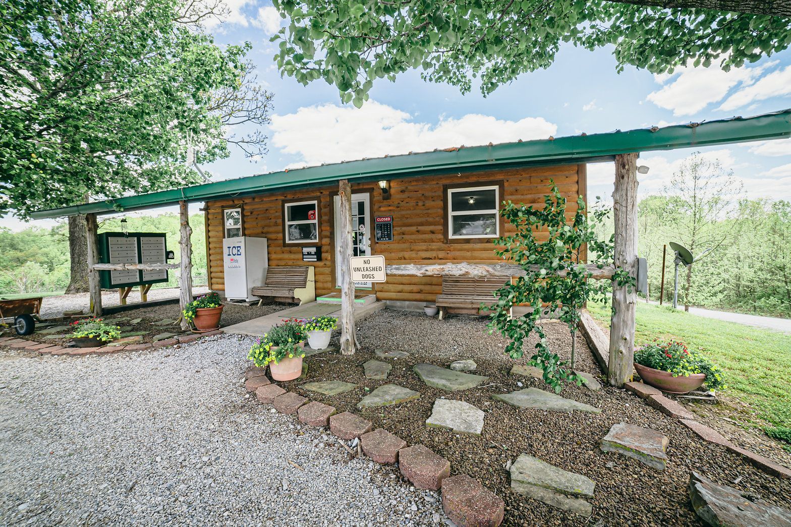 A log cabin exterior with a green metal roof, gravel path, garden beds, and a covered porch area with a white refrigerator.