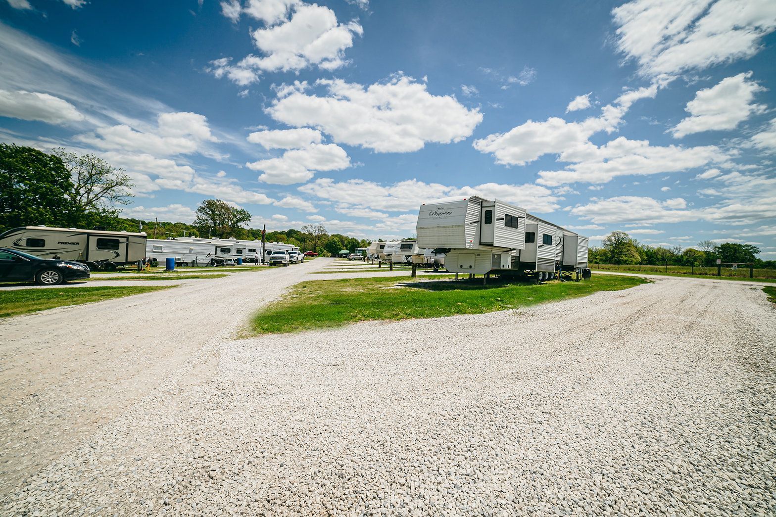 A white fifth-wheel camper parked on a gravel lot at a sunny RV campground with blue sky and scattered clouds.