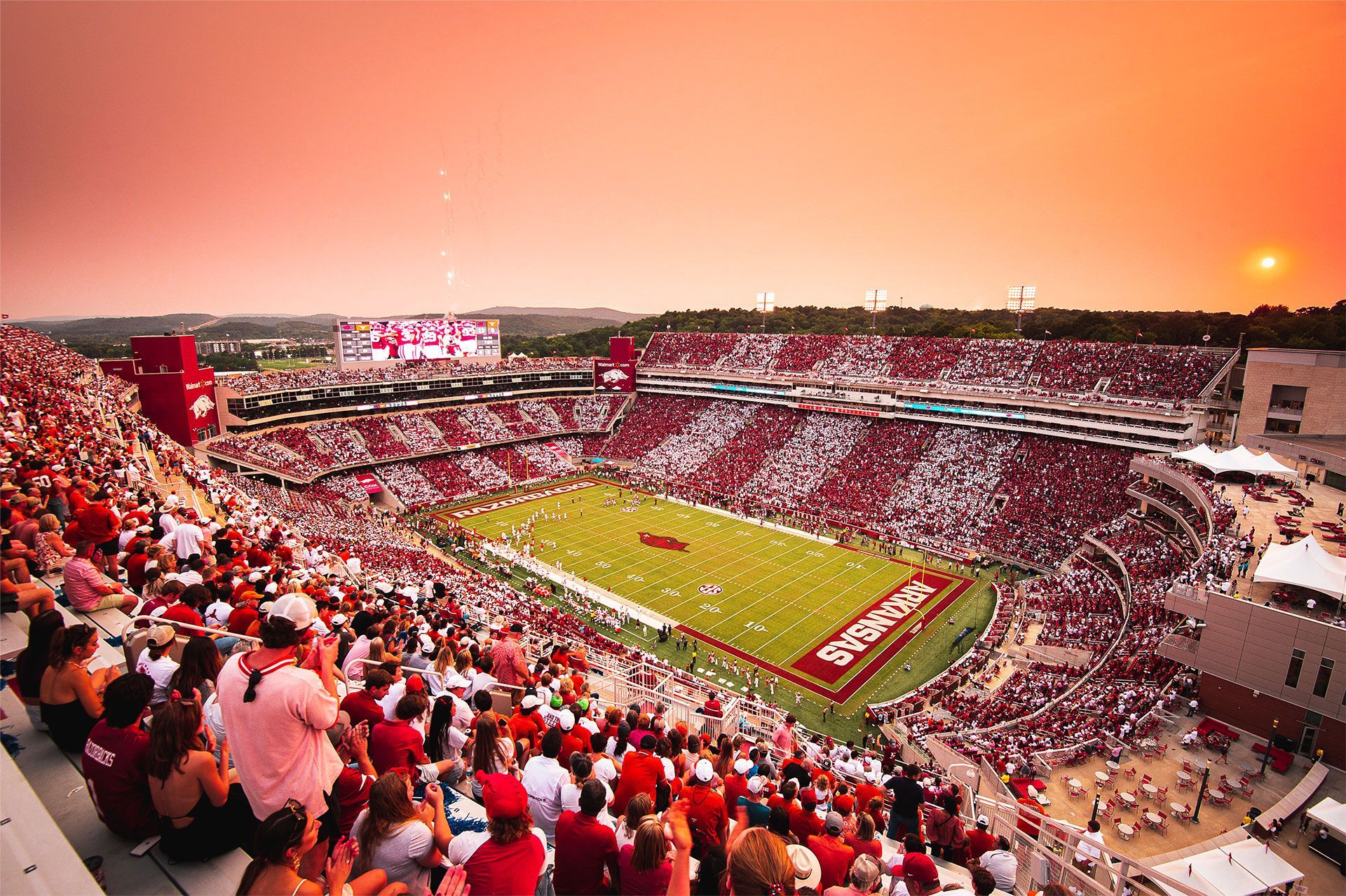 Aerial view of a packed football stadium during sunset, with fans wearing red and white in a vibrant, sunlit setting.
