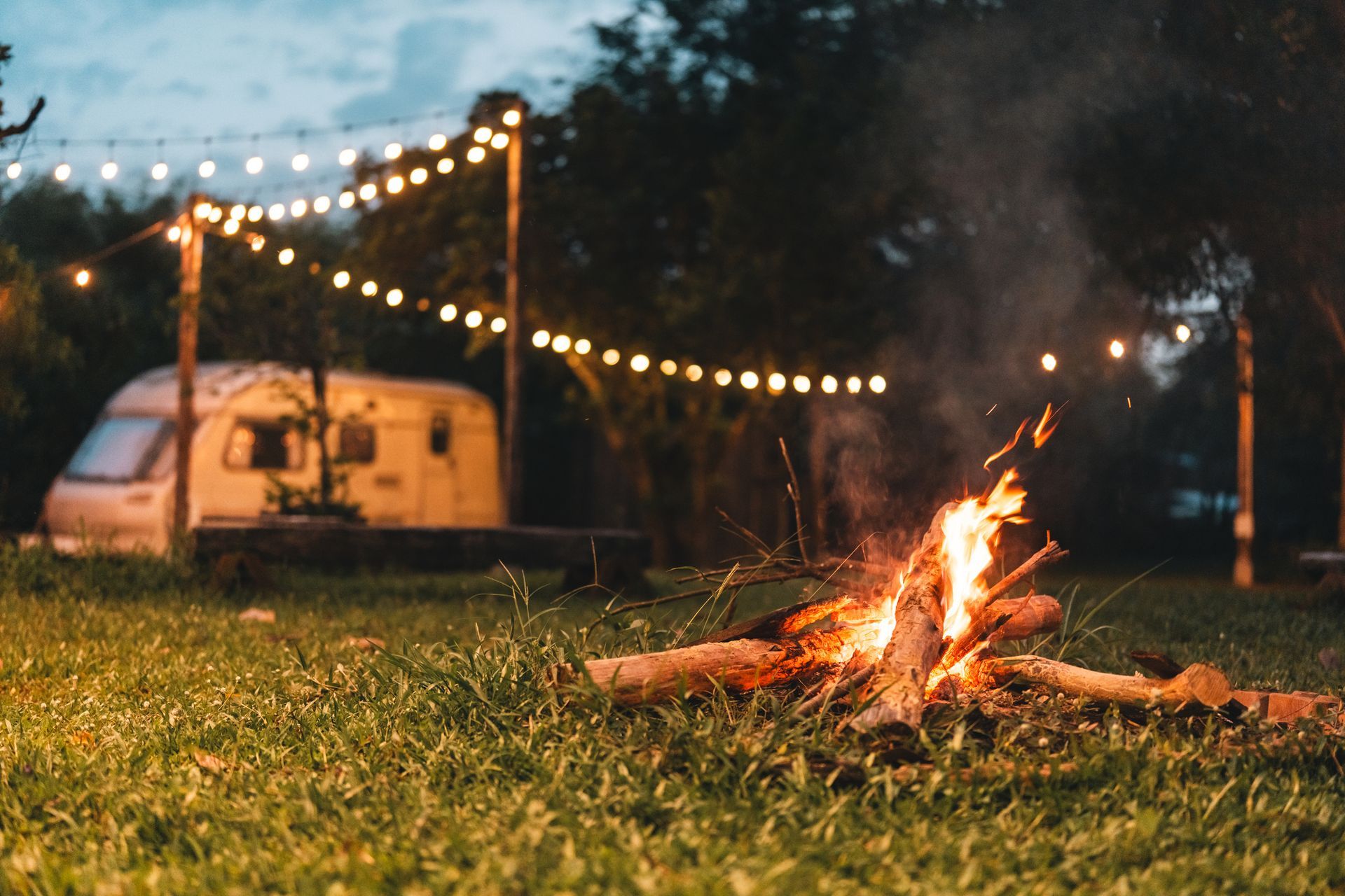 A small campfire burns on grass beside a lit camper and string lights at dusk.