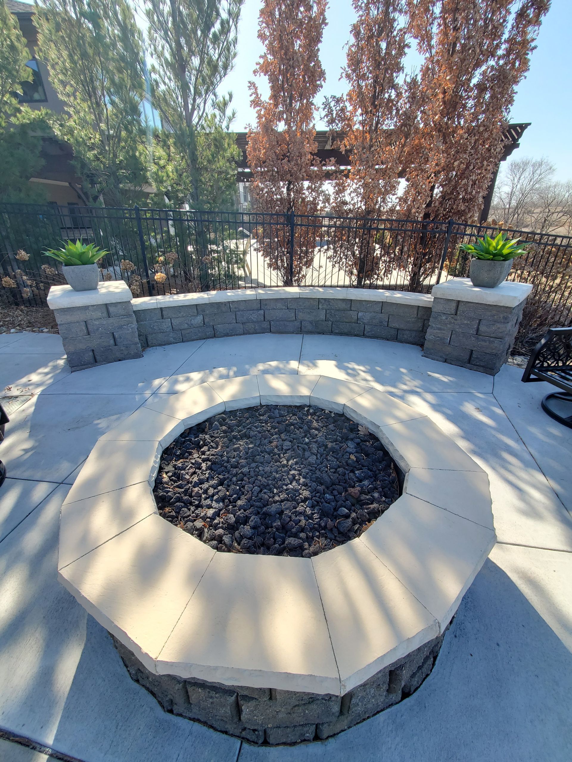 Circular stone fire pit with seating, potted plants, and trees in a backyard setting.