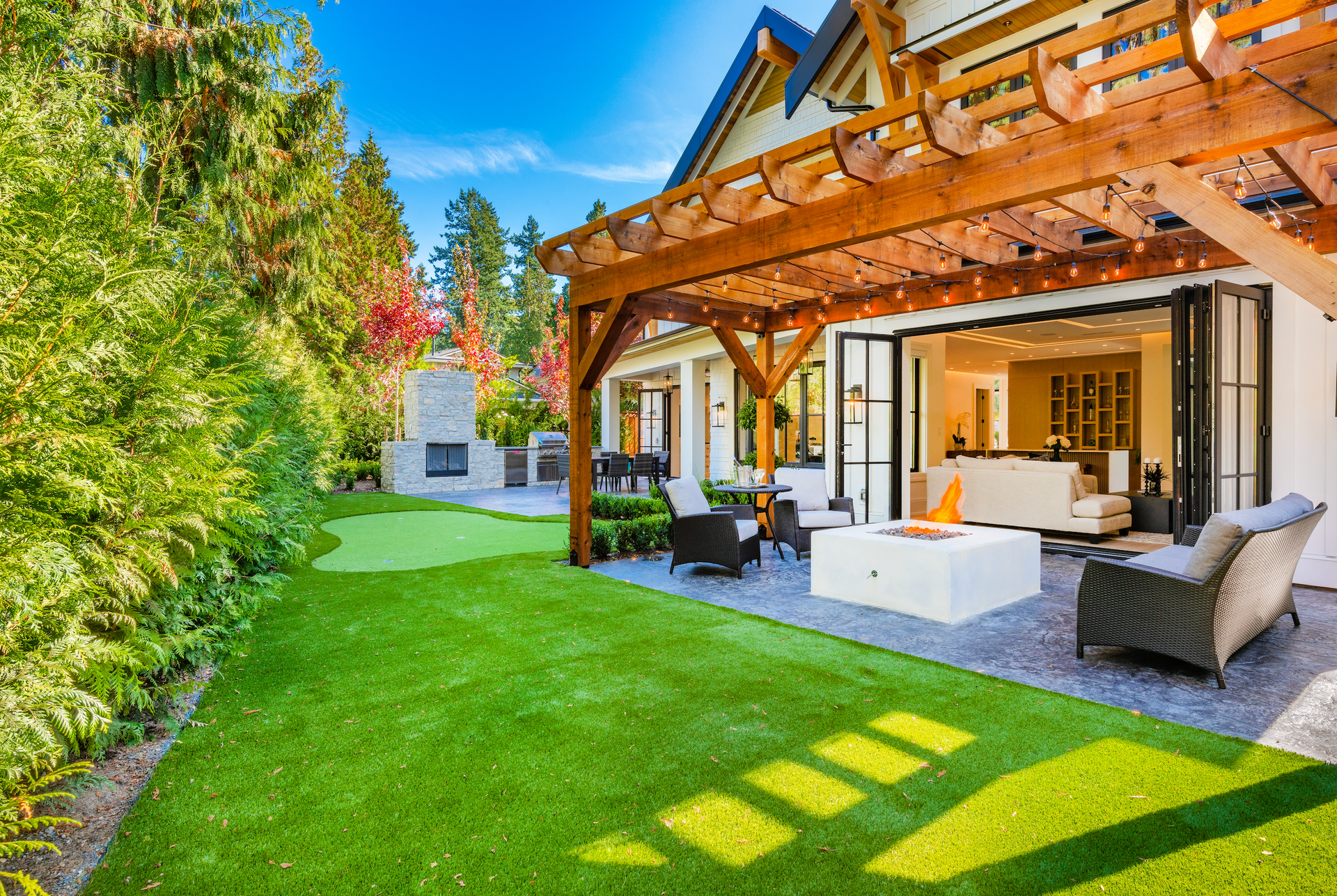 Lush backyard with artificial grass, patio, and open doors to living room. Wooden pergola overhead, fire pit, and seating.
