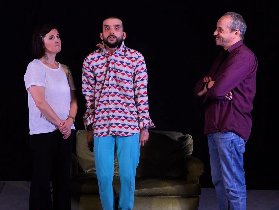 Trois personnes se tiennent debout dans un studio sombre, avec un canapé derrière elles et un éclairage violet.