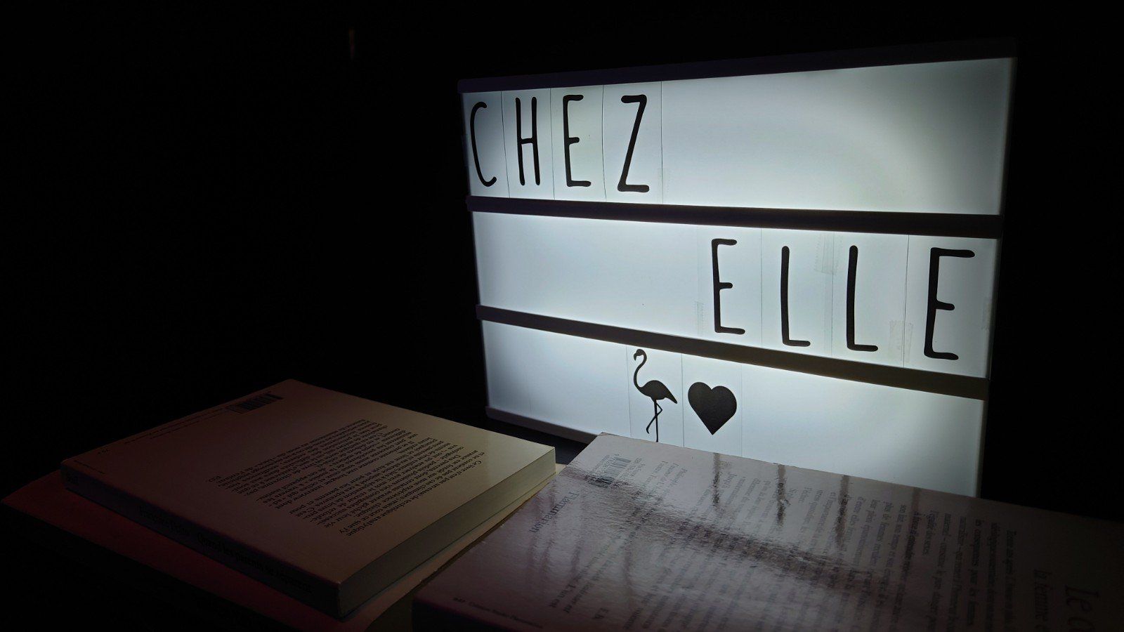 Panneau lumineux indiquant « CHEZ ELLE » à côté d'un livre sur une table dans une pièce sombre