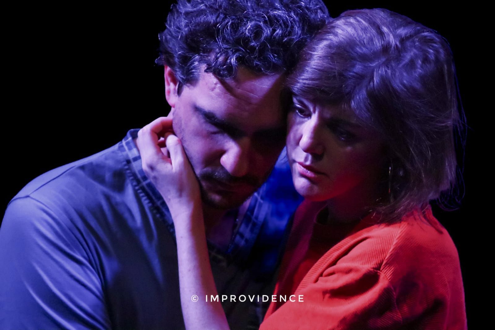Deux personnes enlacées sous une lumière bleu-violet, l'une vêtue d'un haut rouge et l'autre d'une chemise bleue, toutes deux affichant une mine sombre