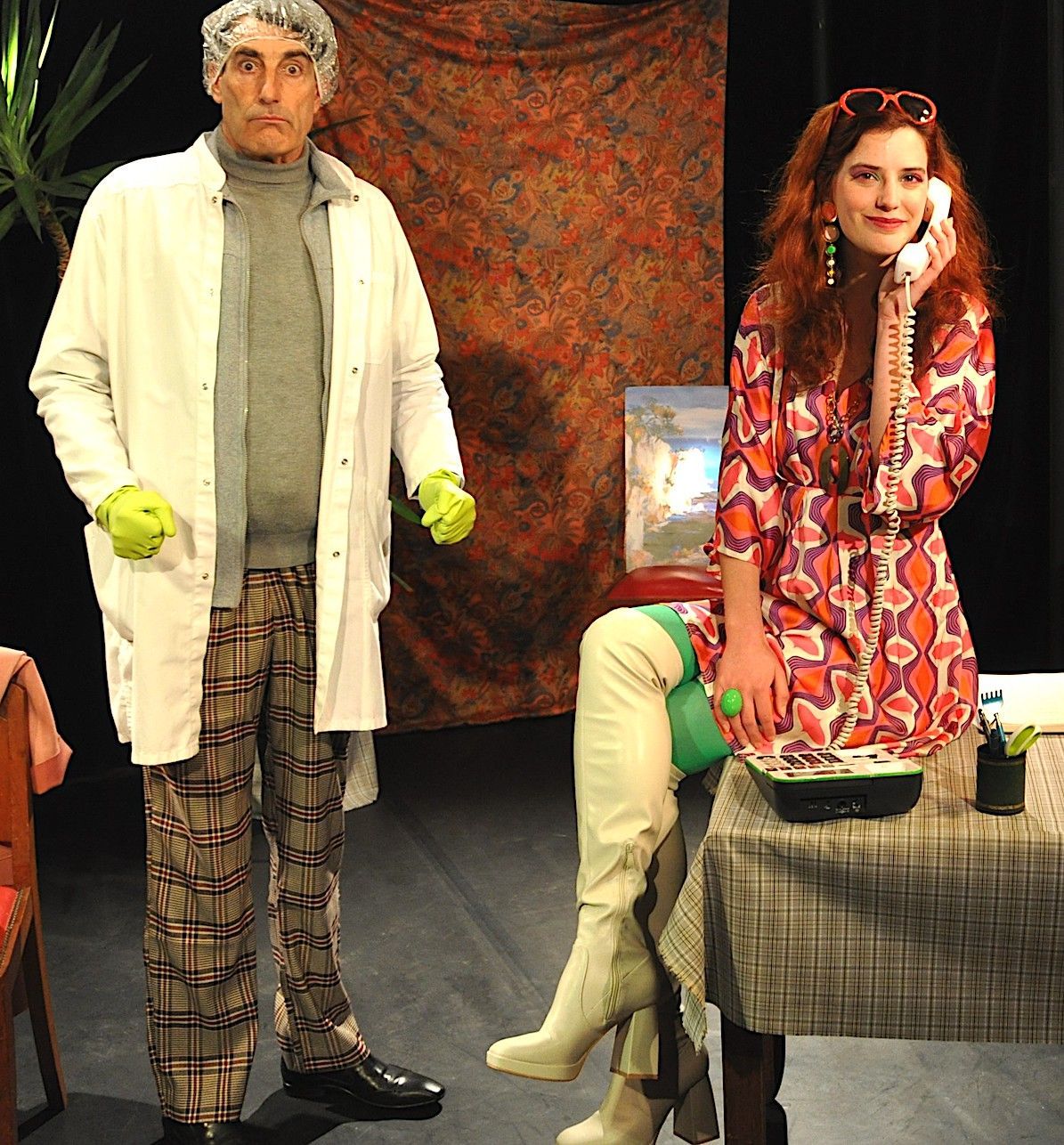 Deux personnes costumées sur scène, l'une debout en blouse blanche et pantalon à carreaux, l'autre assise en robe rouge.