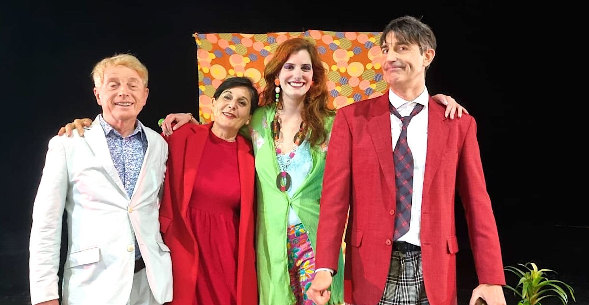 Quatre personnes en costumes colorés se tiennent sur scène, souriantes et posant devant un fond noir.
