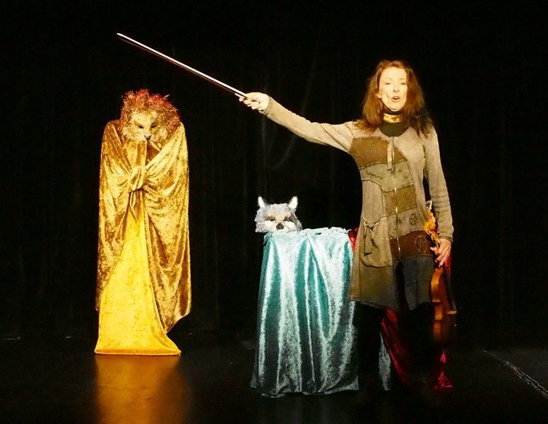 Scène de théâtre avec un personnage de magicien, une femme tenant une baguette magique et un chat sur un piédestal drapé.