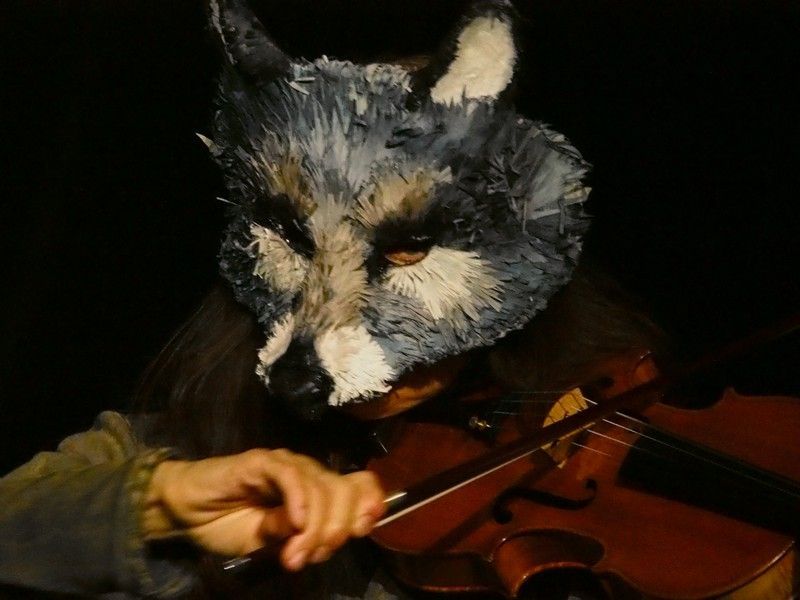 Une personne portant un masque de loup joue du violon dans un décor faiblement éclairé.