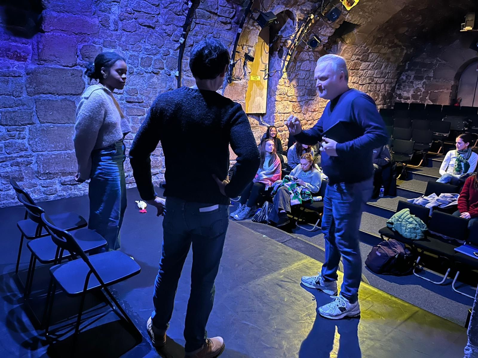Un petit groupe discute dans une cave en pierre éclairée de bleu, avec des chaises pliantes et un arrangement floral.