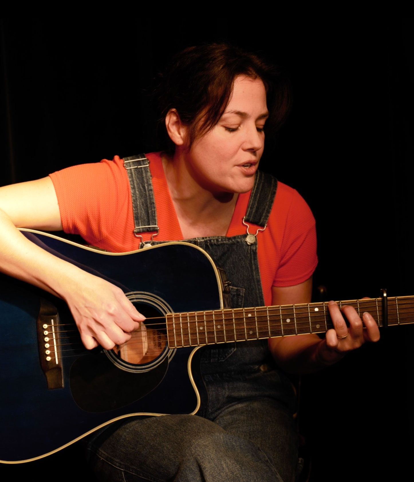 Une femme en chemise orange et salopette joue de la guitare acoustique noire sur une scène sombre