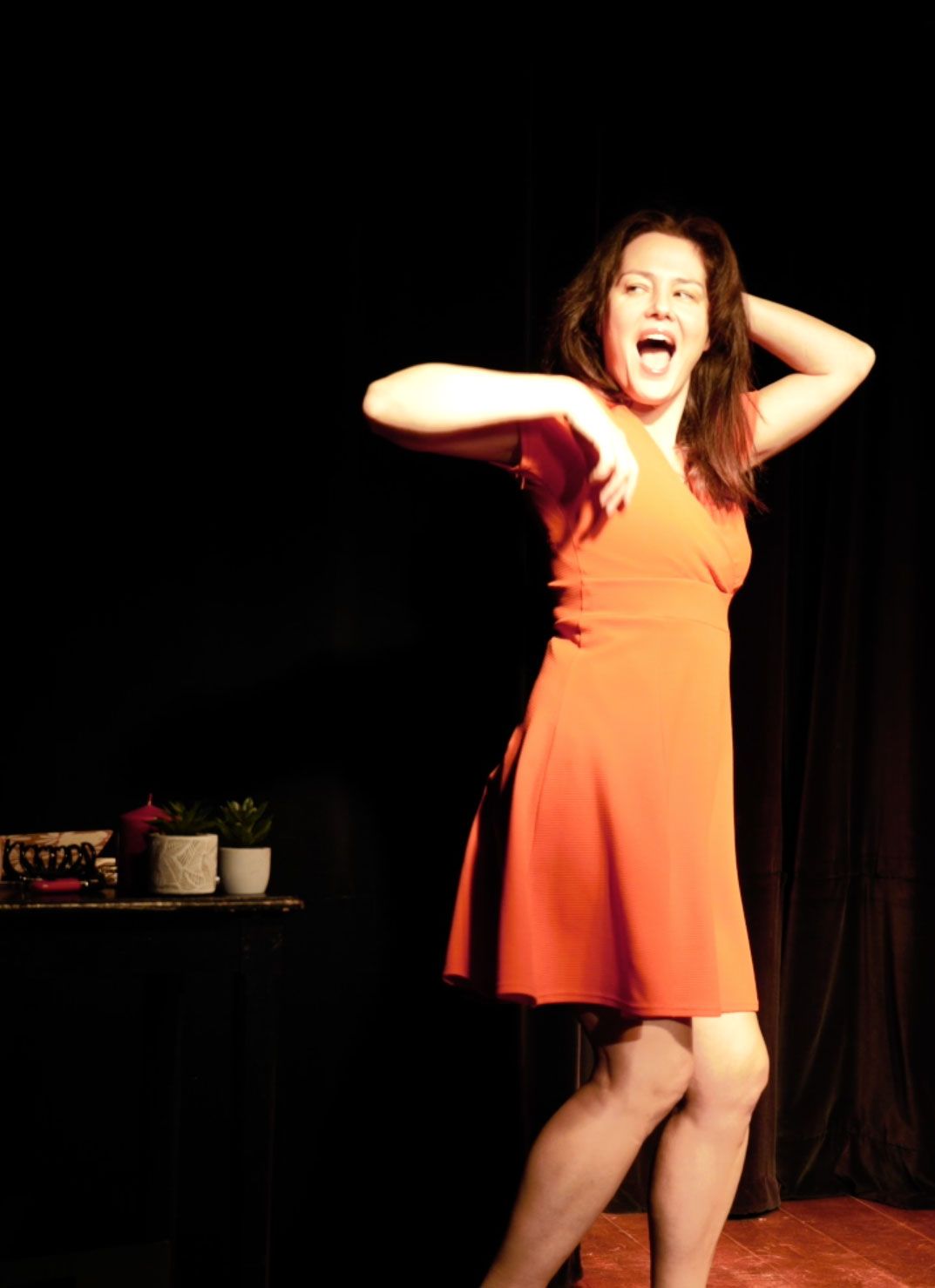 Une femme en robe orange pose sur scène, les mains derrière la tête, sous les projecteurs.