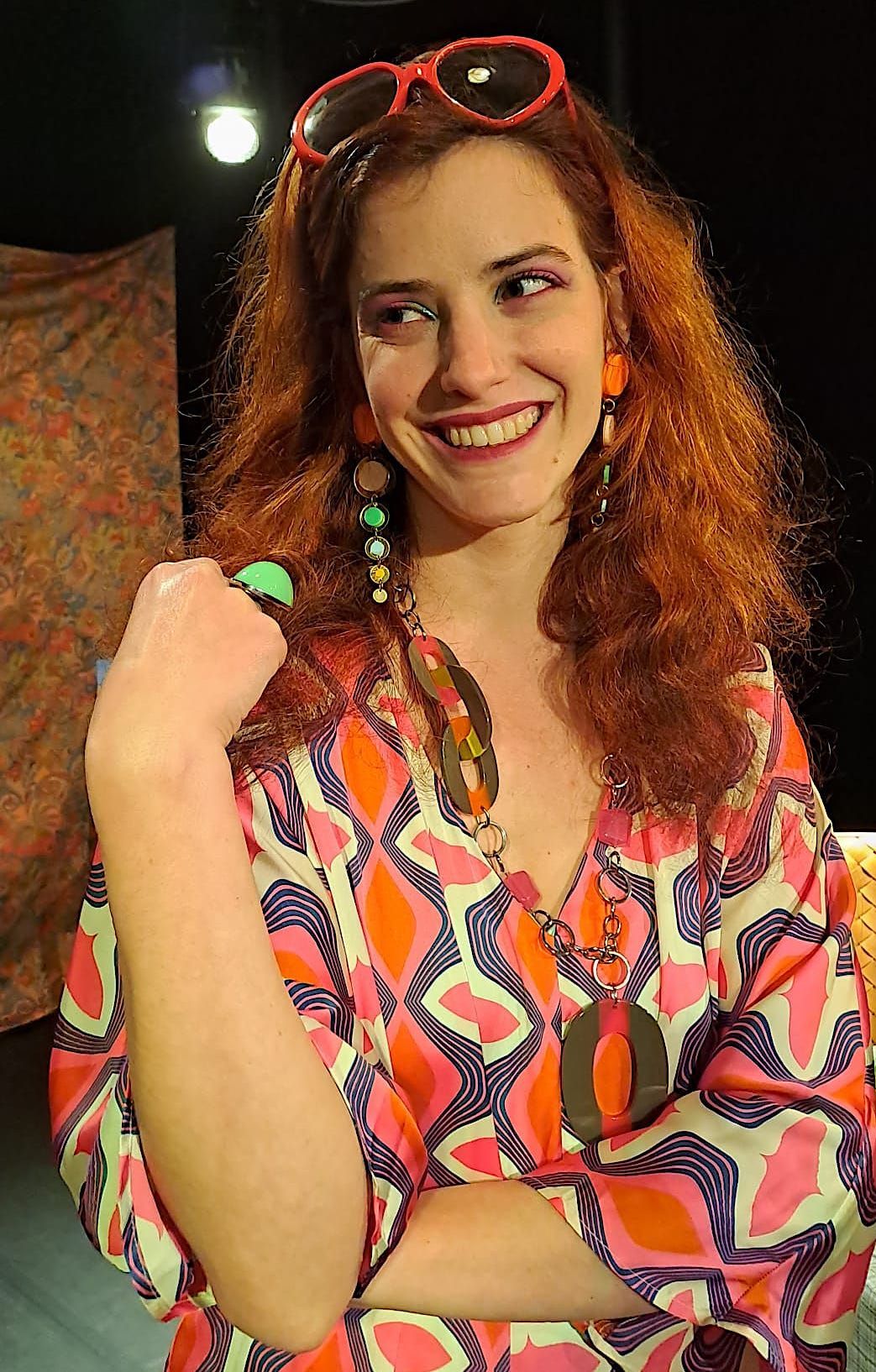 Personne souriante vêtue d'une robe à motifs colorés, les cheveux roux et portant des lunettes de soleil, se détachant sur un fond sombre.