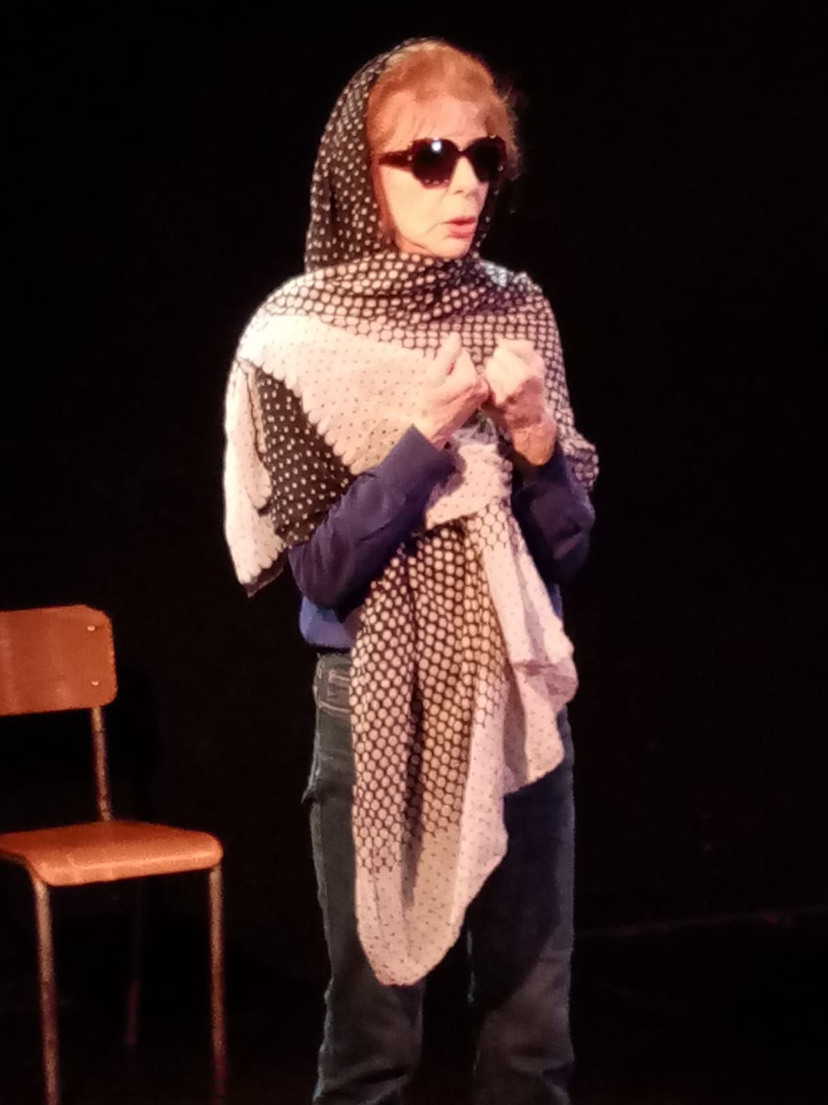 Une personne sur scène portant des lunettes de soleil et une écharpe à motifs, à côté d'une chaise en bois.