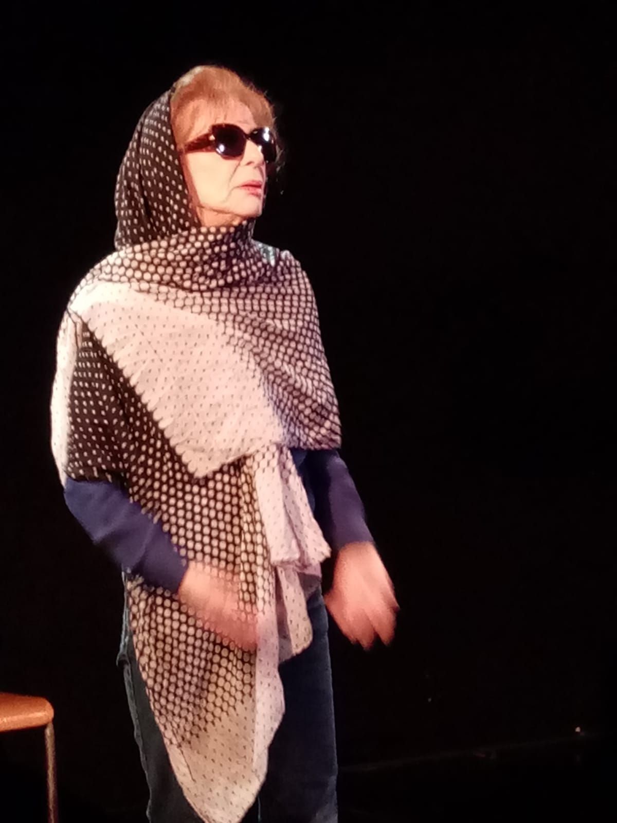 Une femme sur une scène sombre, portant des lunettes de soleil, un foulard à motifs et un châle léger, se tient près d'une chaise.