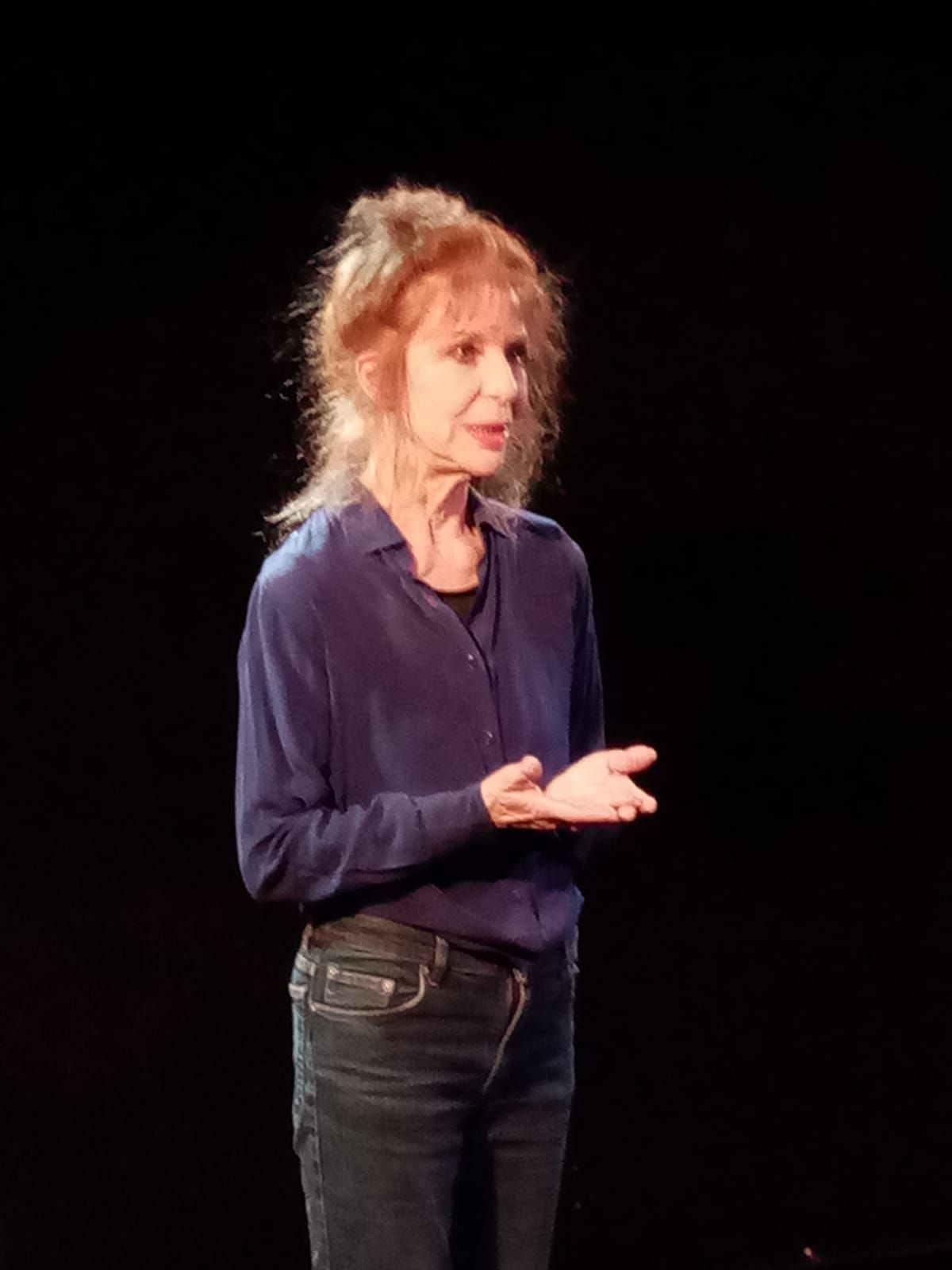 Une femme en chemise bleu foncé et jean noir prend la parole sur une scène sombre.