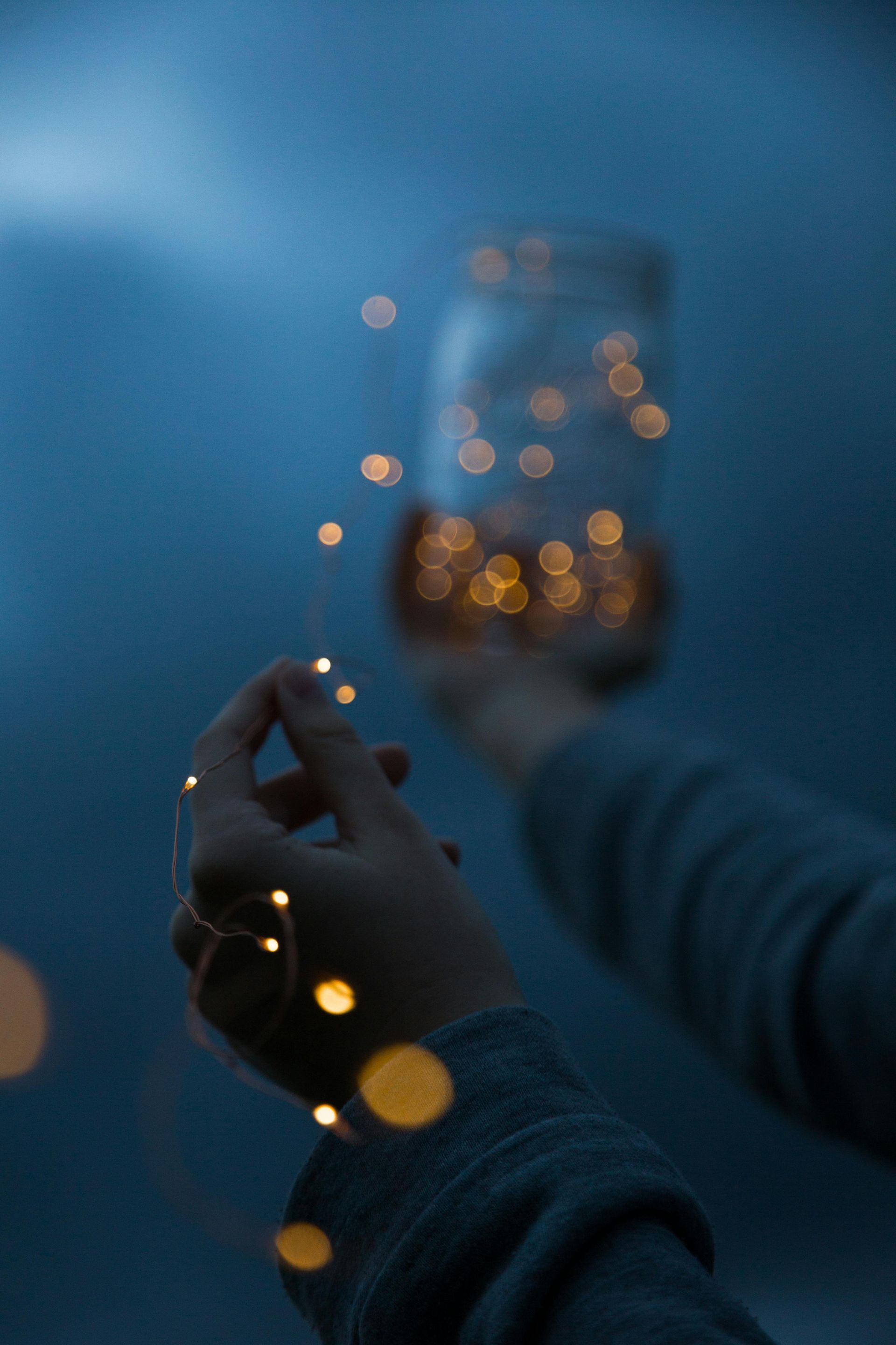 Des mains tenant une guirlande lumineuse aux tons chauds dans une pièce bleuâtre tamisée, avec un bocal à la lueur floue en arrière-plan.