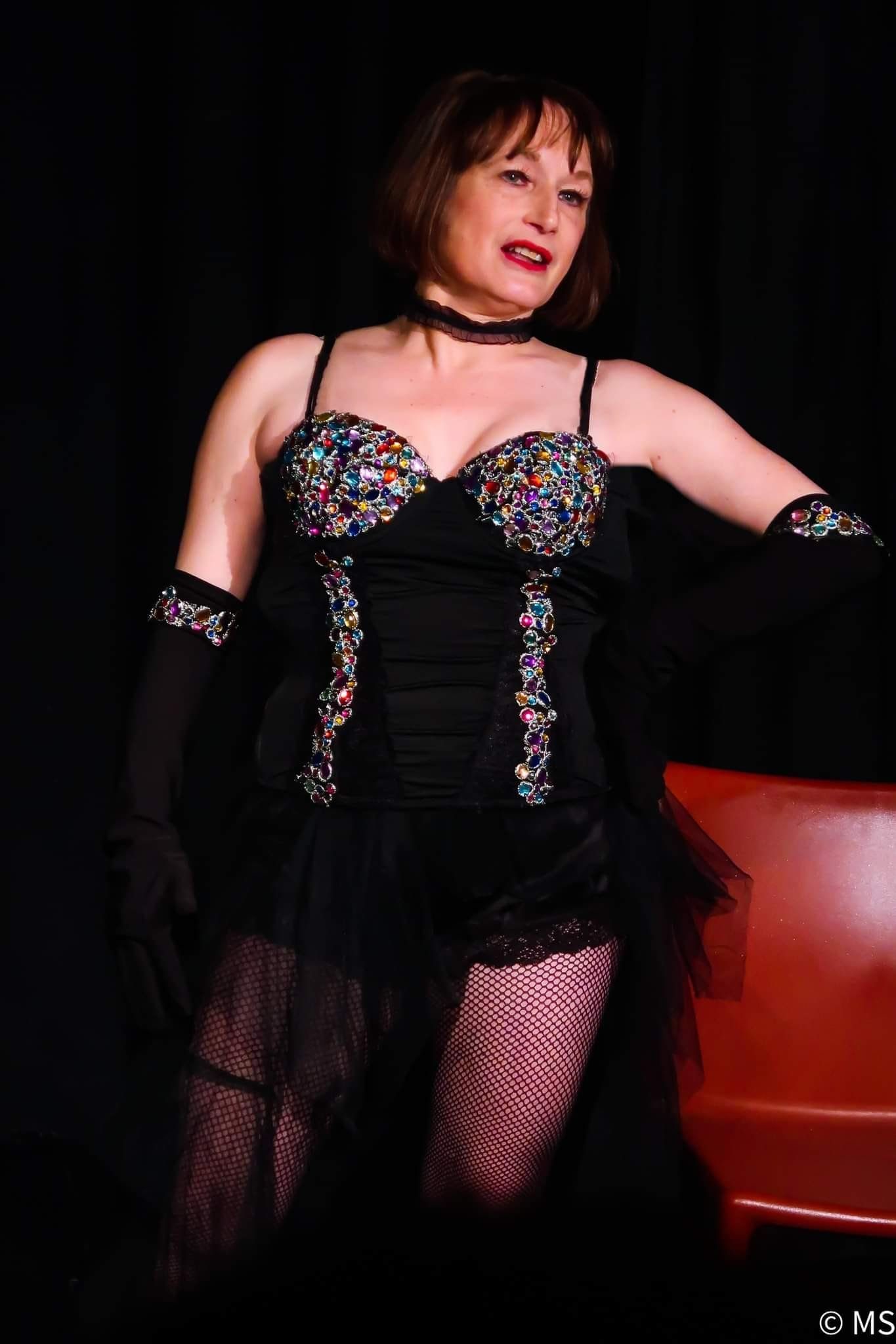 Femme en corset noir et argent posant sur fond sombre à côté d'une chaise rouge