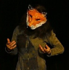 Personne en veste sombre portant un masque de renard orange sur fond noir, les mains levées.
