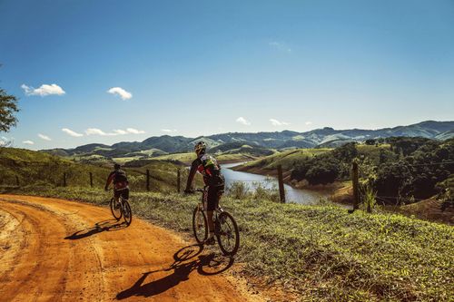 Two cyclists ride mountain bikes along a sunny dirt path overlooking a river in a hilly landscape.