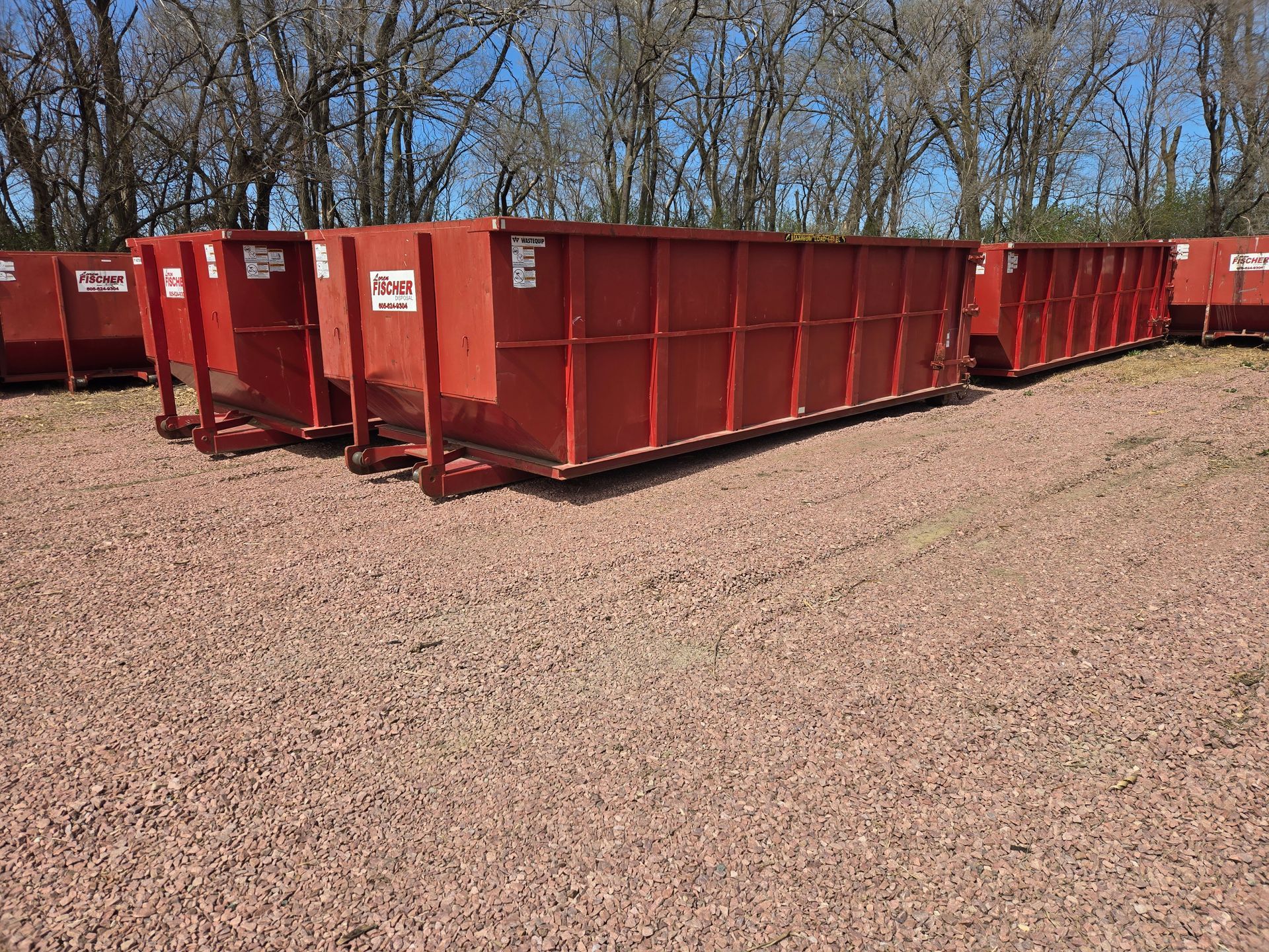 A large green dumpster truck parked on a gravel lot near a chain-link fence, viewed from the rear.
