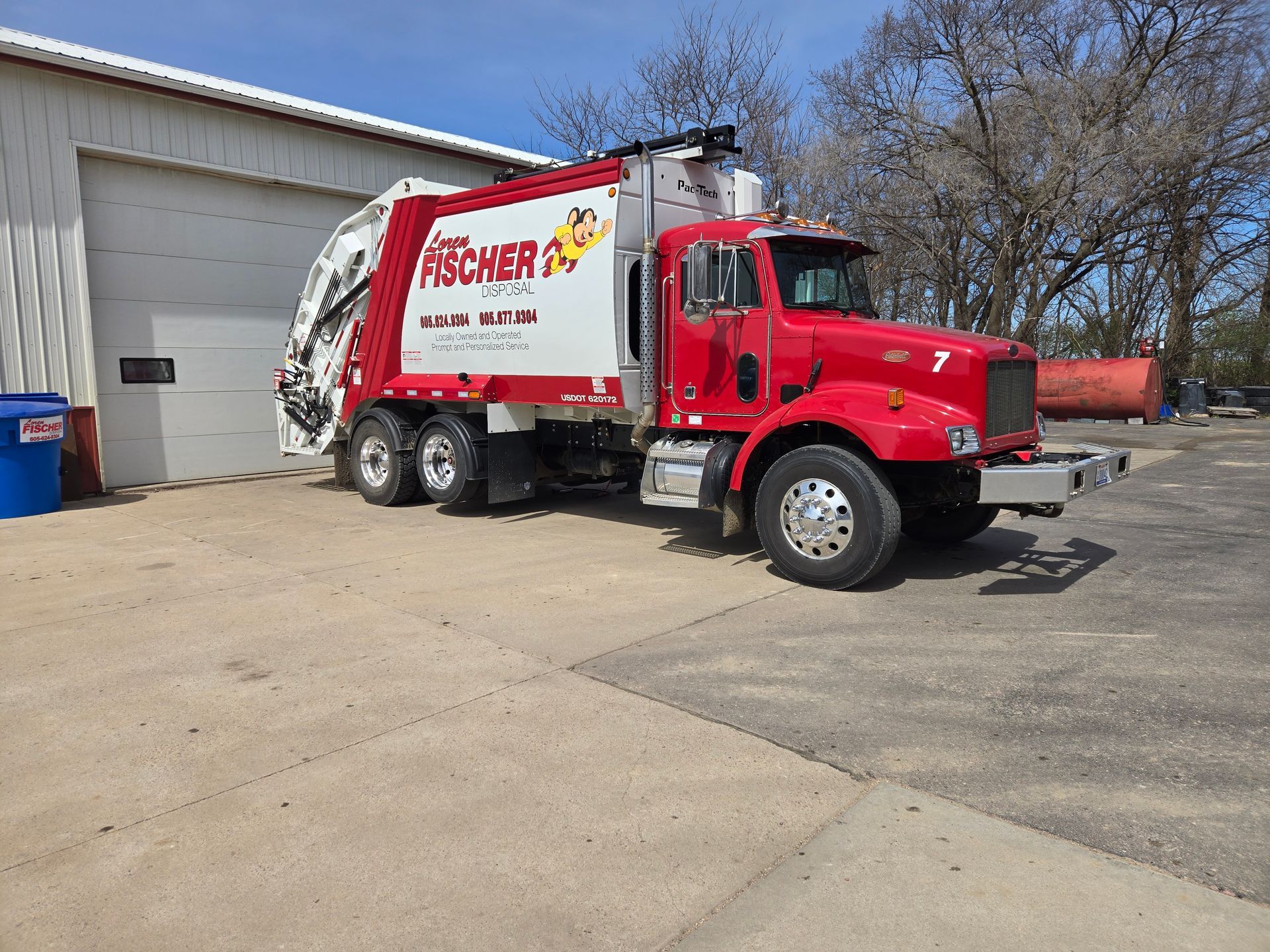 A bright red crane truck lifts a metal container from a recycling station on a roadside.