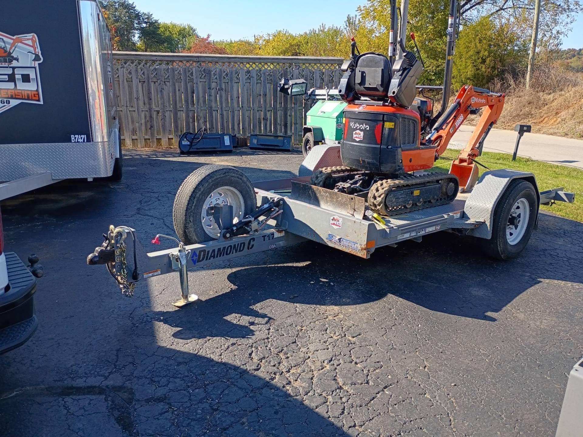 An orange mini-excavator is parked on a flatbed trailer outdoors, positioned on an asphalt surface.
