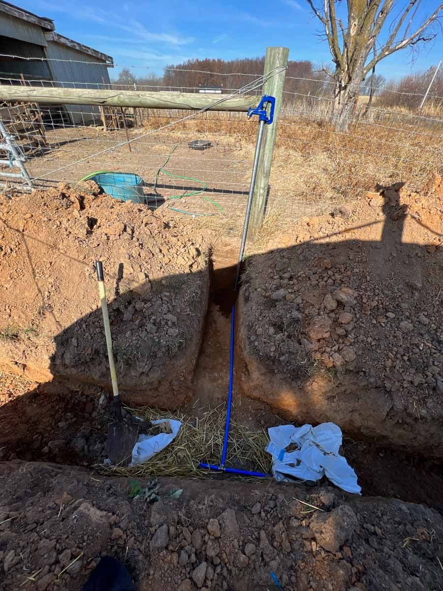 A narrow trench dug in dirt, featuring a shovel and a blue T-handle tool, located in front of a wooden fence post.