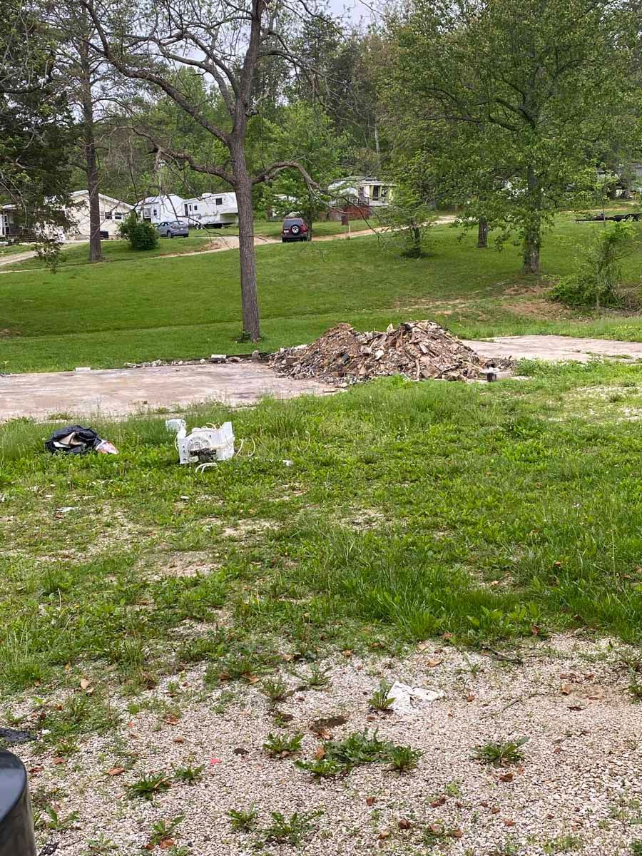 A yard with a gravel driveway, a pile of dry leaves, scattered trash, and houses in the background.