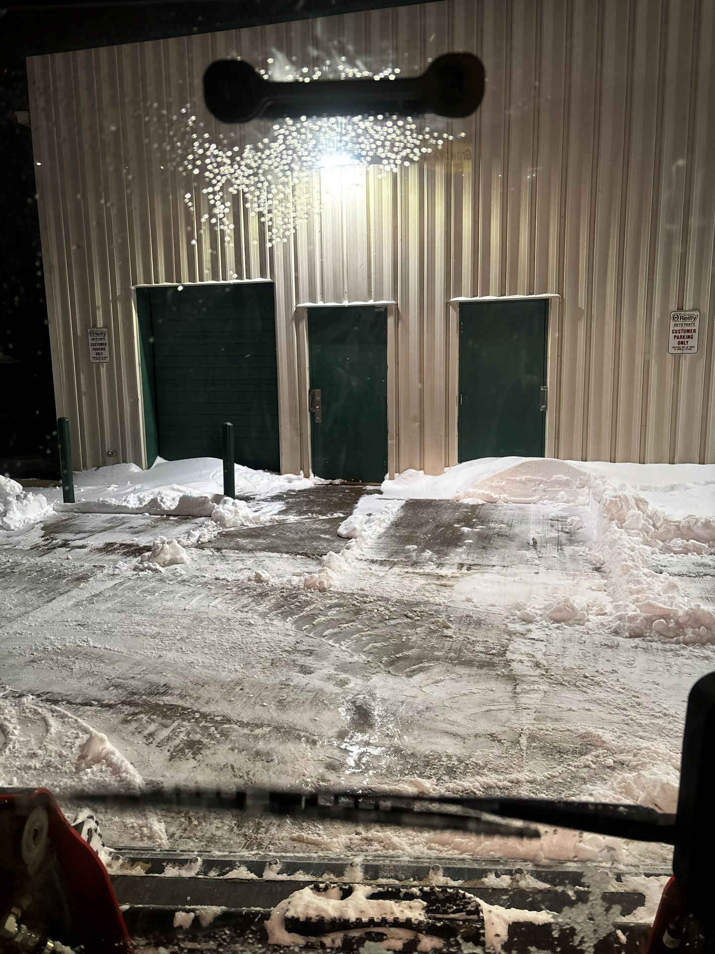 A view from a vehicle cab showing a metal building with three dark green doors surrounded by snow at night.