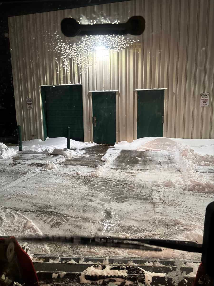 View from a snowplow cab facing a metal building with three green doors after a snowstorm.