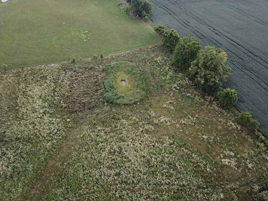 An aerial view of a circular pond surrounded by brush and grasses in a field next to a dark plowed agricultural plot.