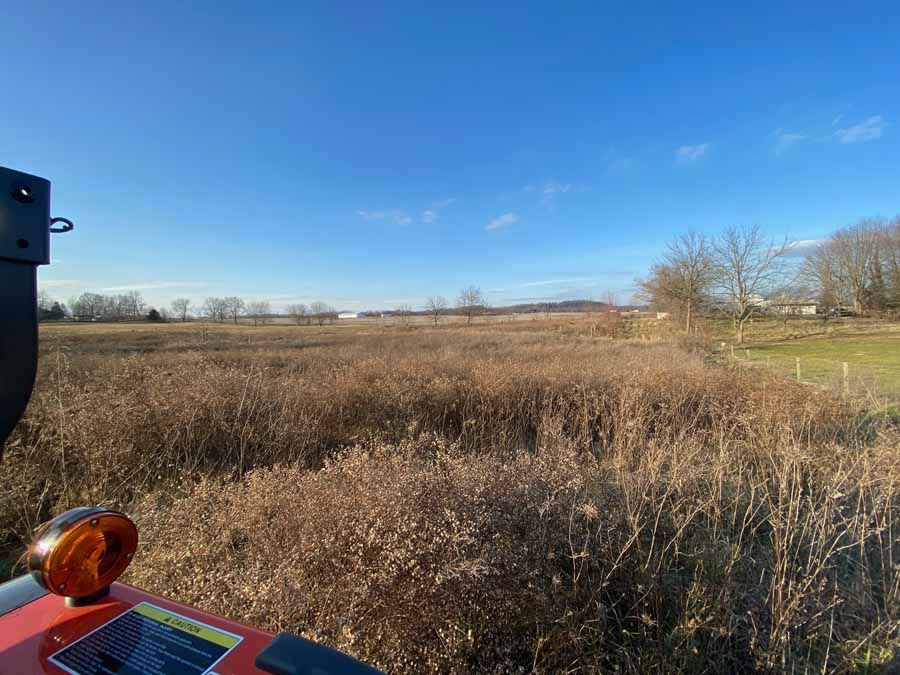 View from the seat of a tractor looking out over a dry, brush-filled field under a clear blue sky.