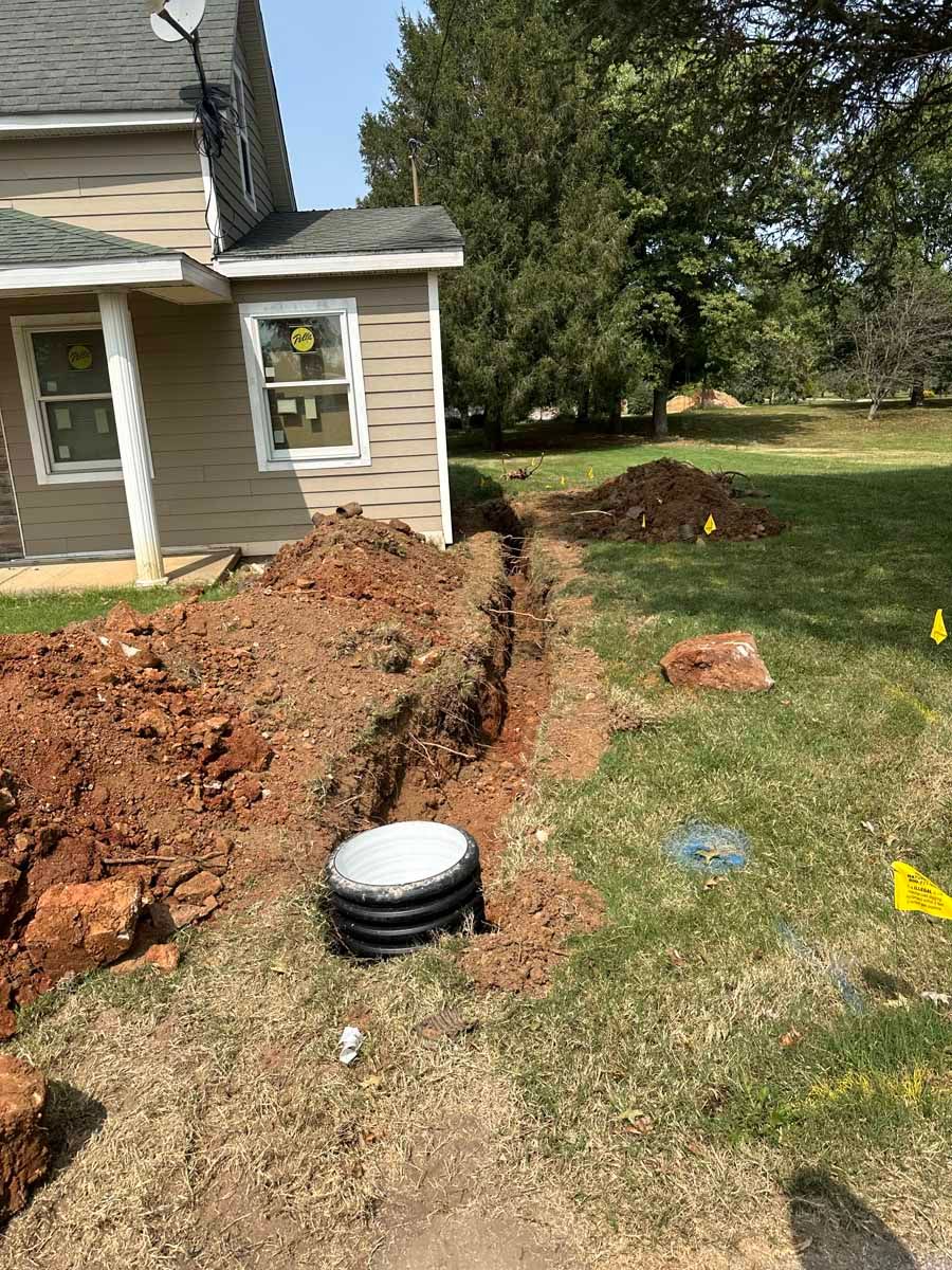 A deep trench and a black drainage basin sit in the grass next to the side of a beige house, with piles of dirt nearby.