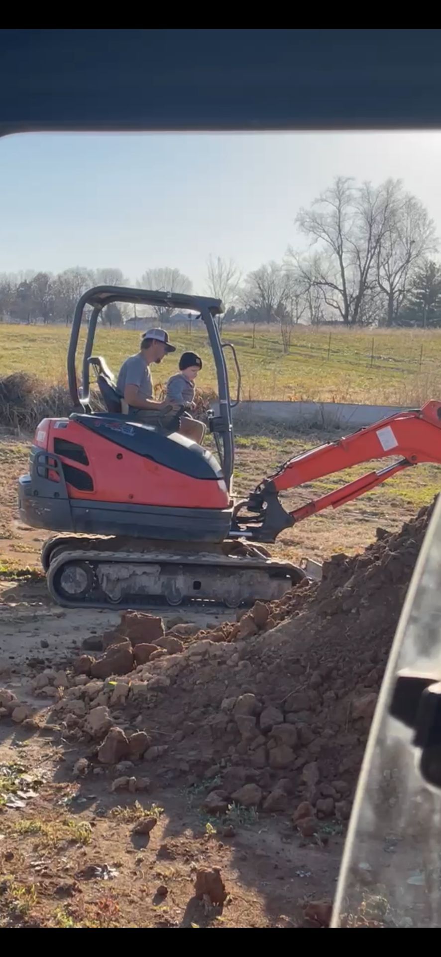 An adult and child sit together on a small orange excavator, operating the machine in a sunny, open dirt lot.