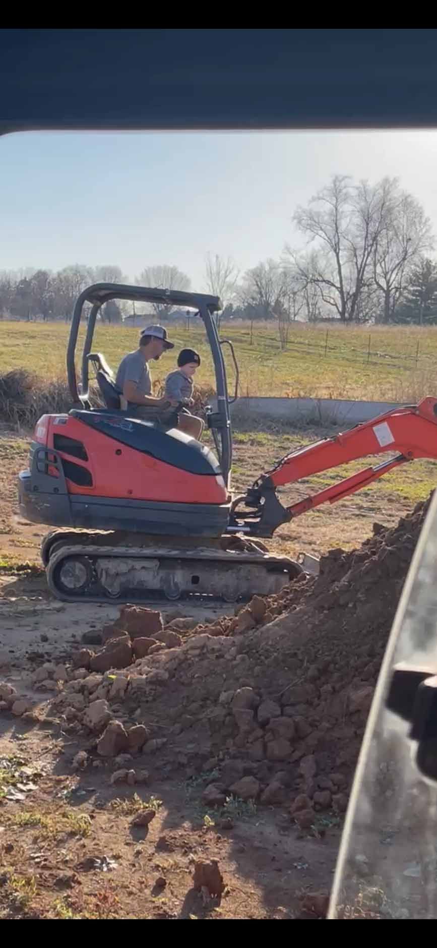 A person and a child sit together in the seat of a red excavator parked on a dirt lot in a rural setting.
