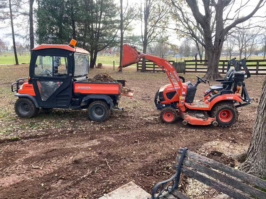 An orange Kubota tractor with a front-loader dumps dirt into an orange Kubota utility vehicle in a grassy, wooded yard.