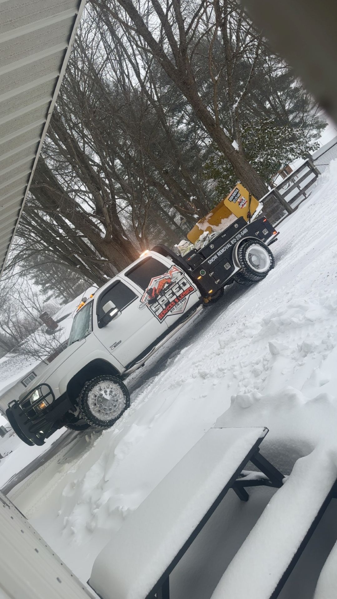 A white pickup truck with a salt spreader on its flatbed parked on a snowy, residential lawn near a wooden picnic table.