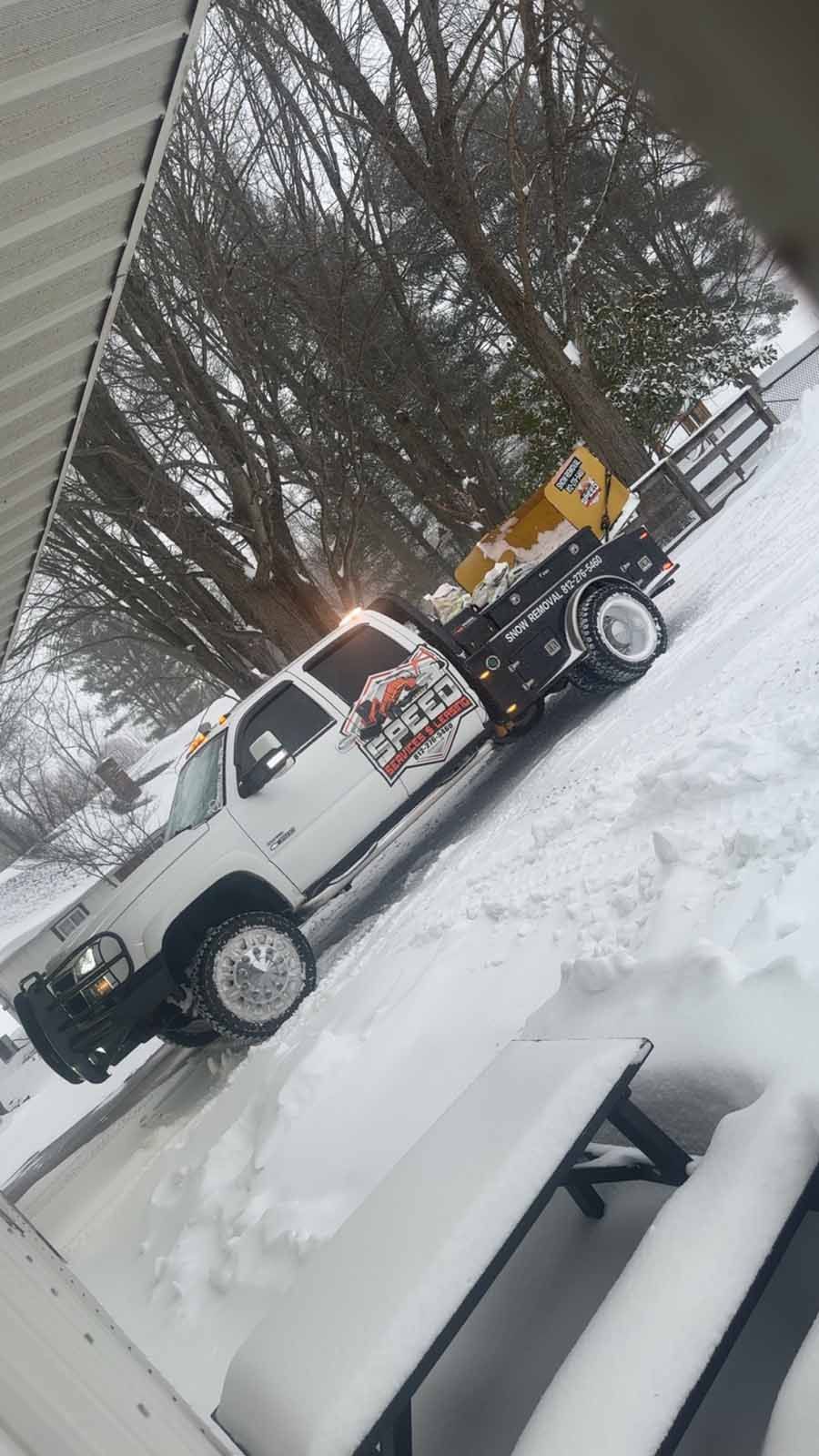 A white flatbed truck with a yellow salt spreader is parked in a snow-covered yard next to a picnic table.
