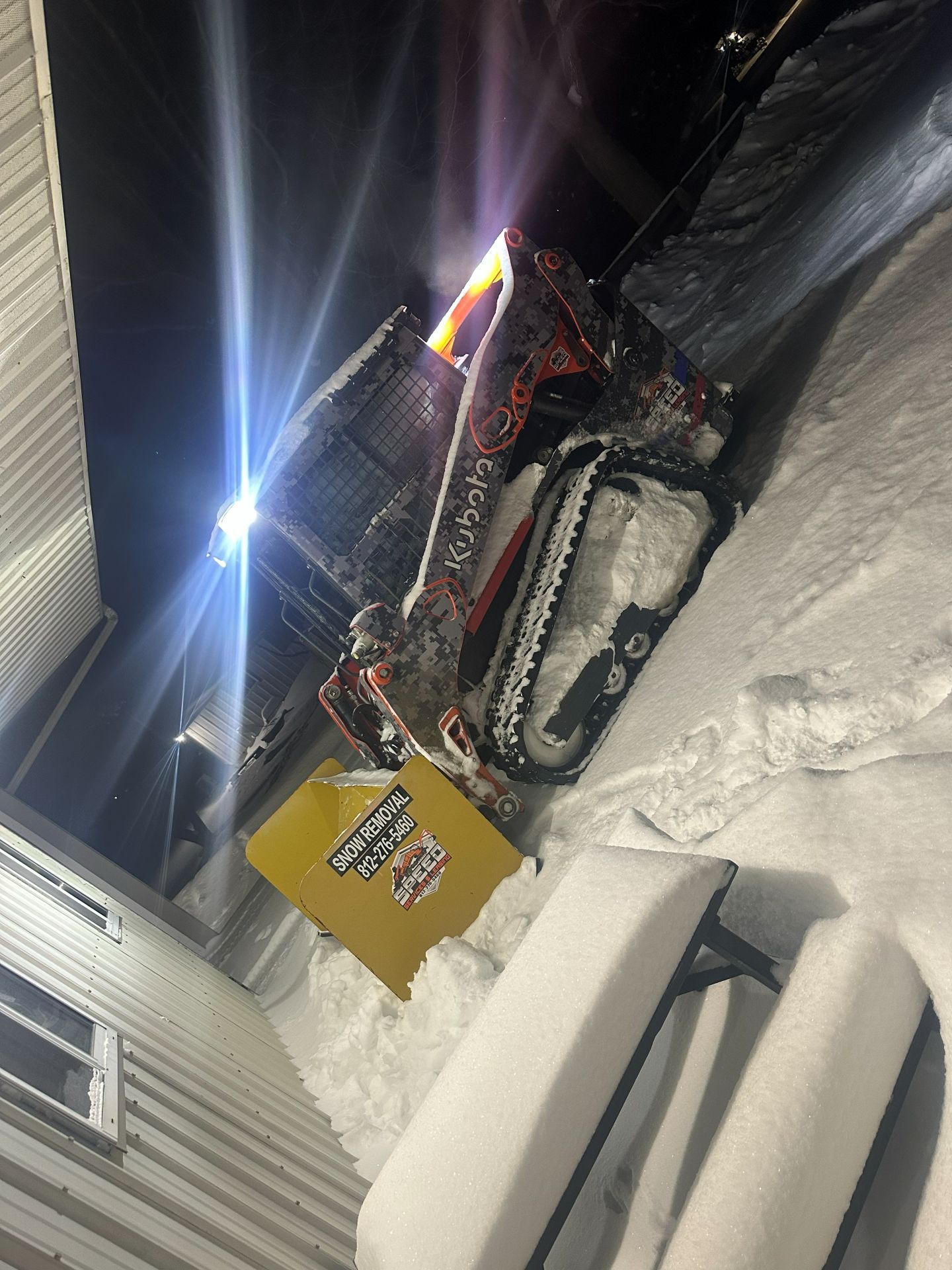 A Kubota tracked skid steer with its lights on, parked next to a yellow metal box in a snow-covered area at night.