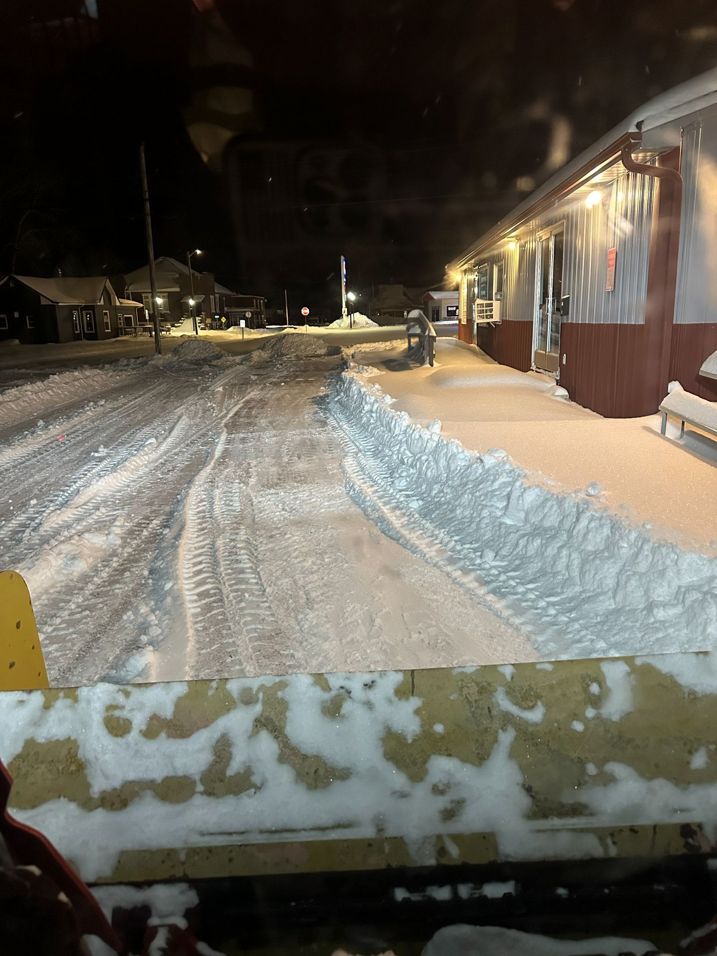 View from a snowplow blade looking at a snow-covered street and building at night.