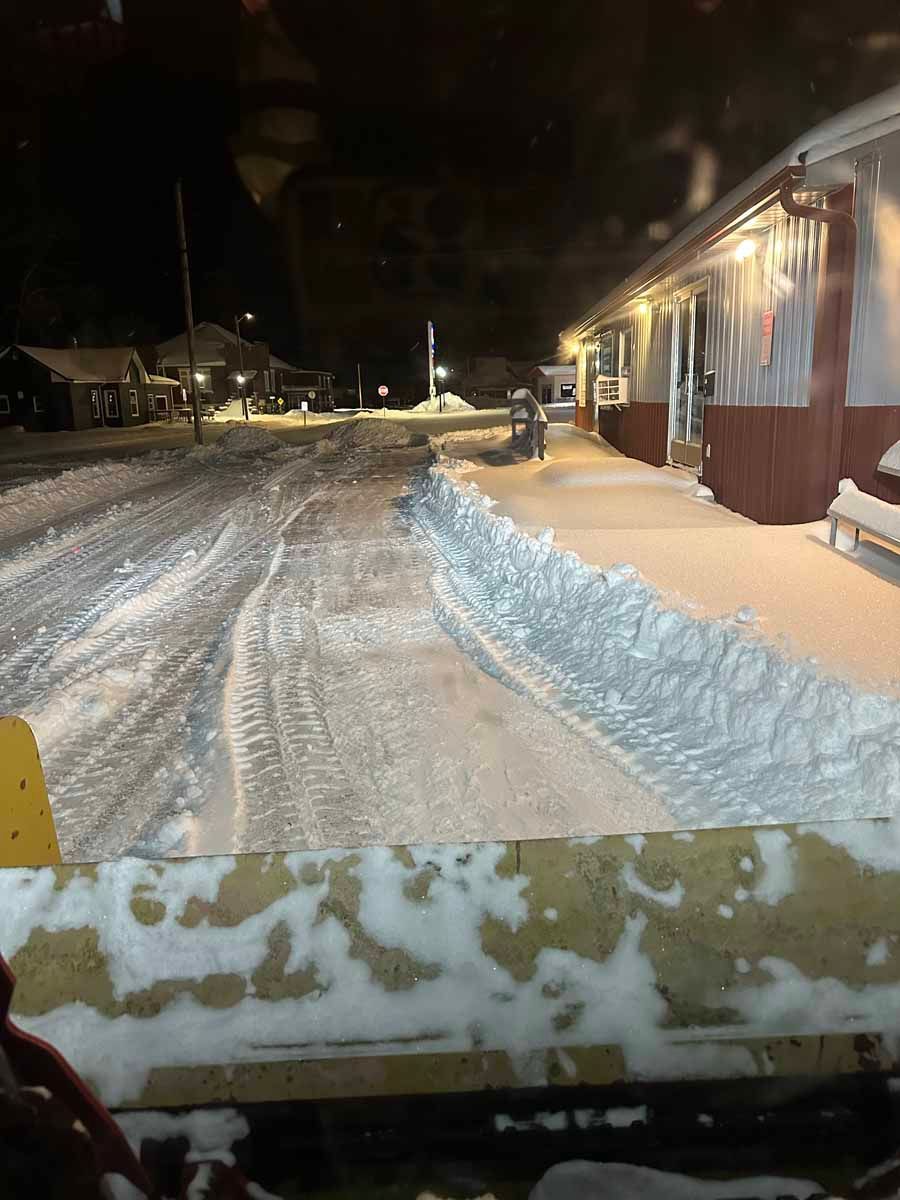 A snowplow blade clears snow in front of a dark, lit building at night after a heavy snowfall.
