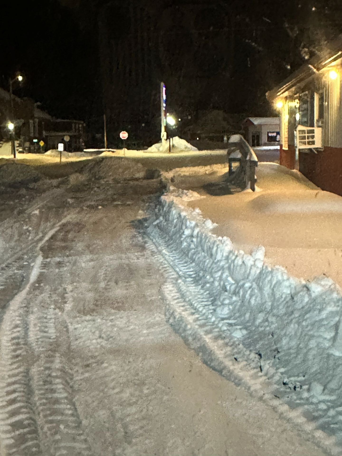 A night scene of a snow-covered street and sidewalk with tire tracks leading toward an intersection near a lit building.