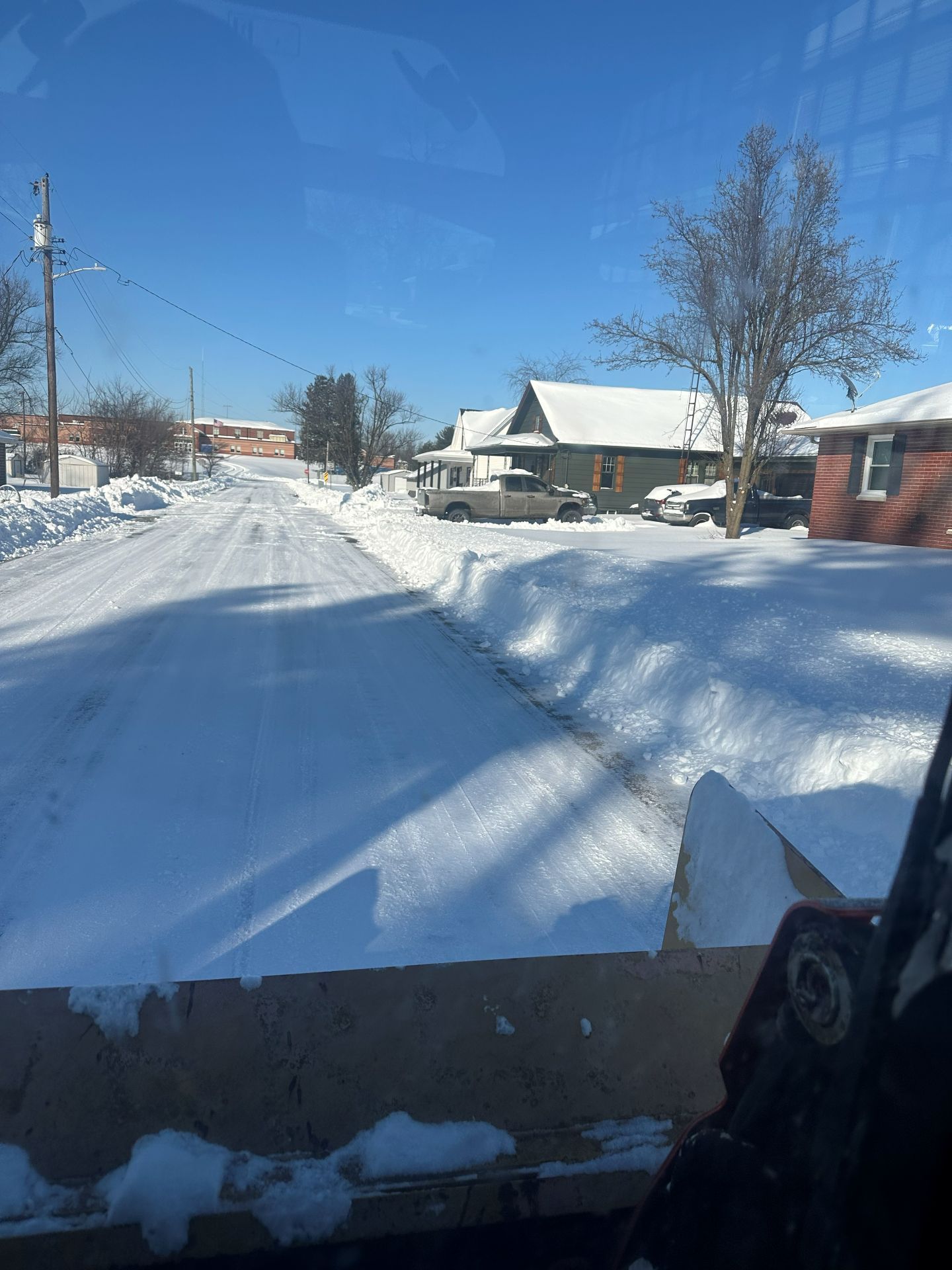 View from a snowplow cab looking down a snow-covered residential street on a sunny day.