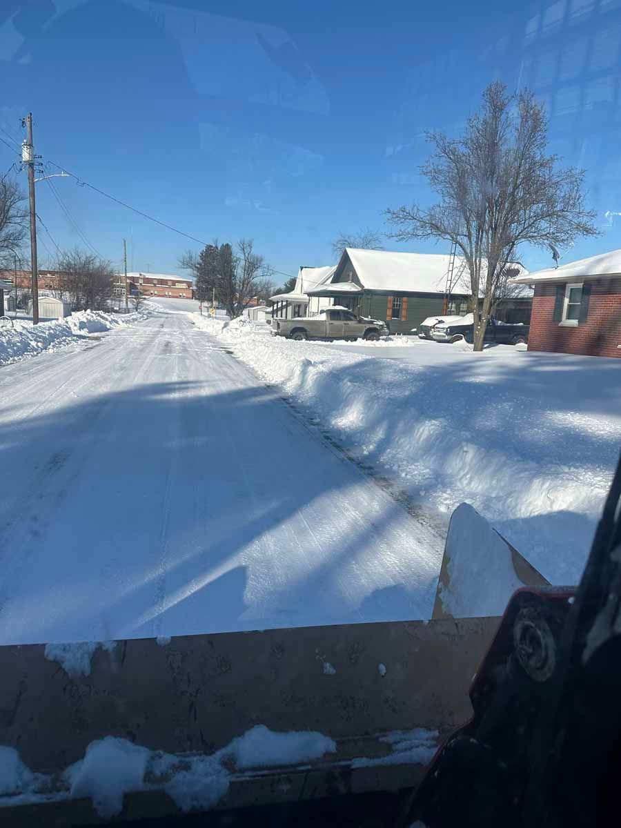 A view from inside a snowplow clearing a snow-covered residential street on a sunny day.