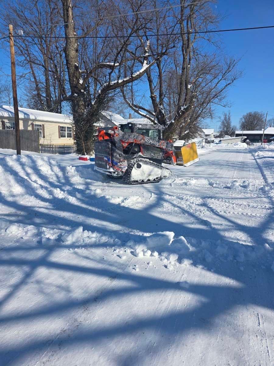 A tracked snow blower clears a snow-covered residential street on a sunny day.