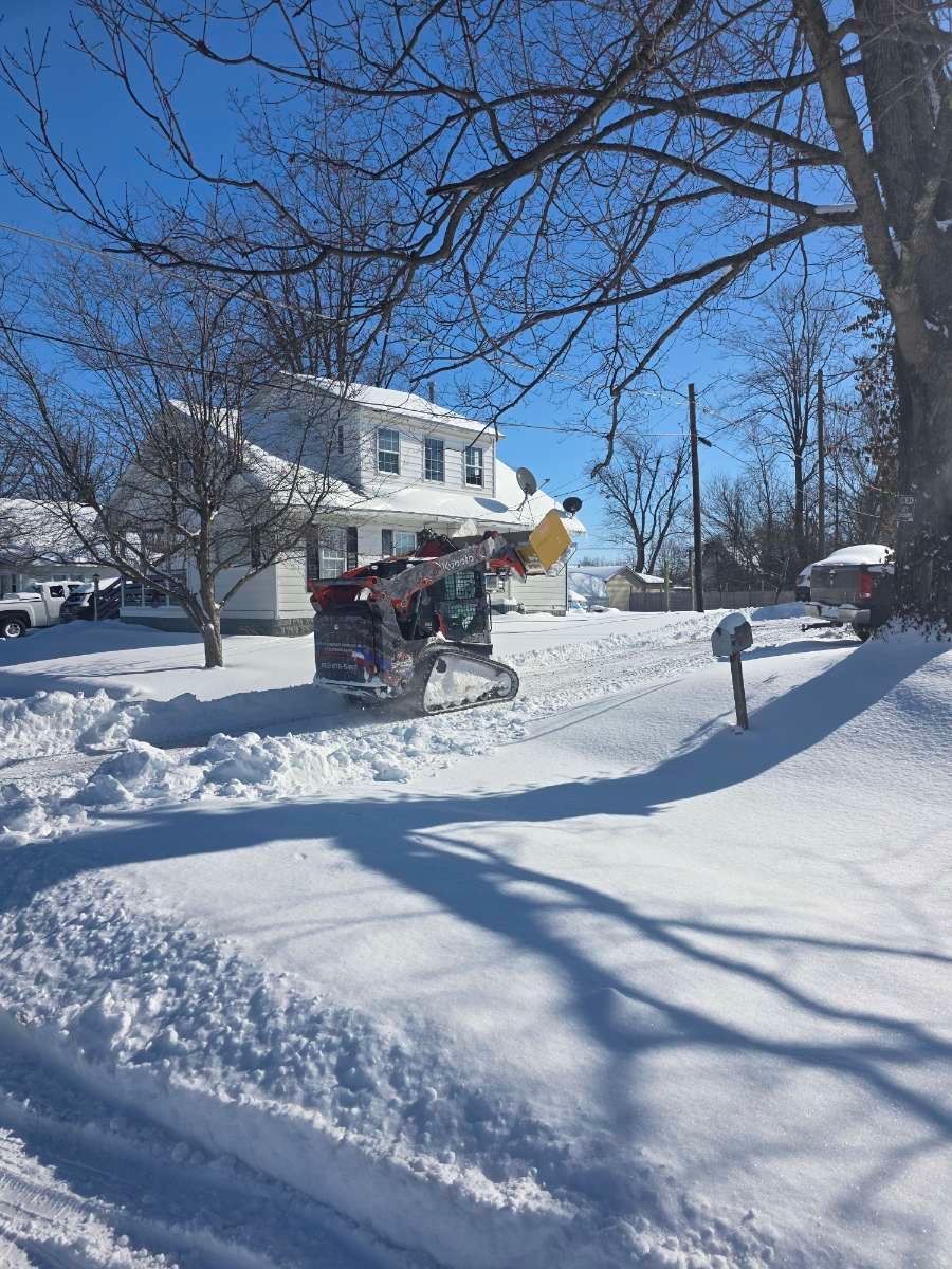 A skid steer loader clears a snow-covered driveway in front of a white house on a bright, sunny winter day.