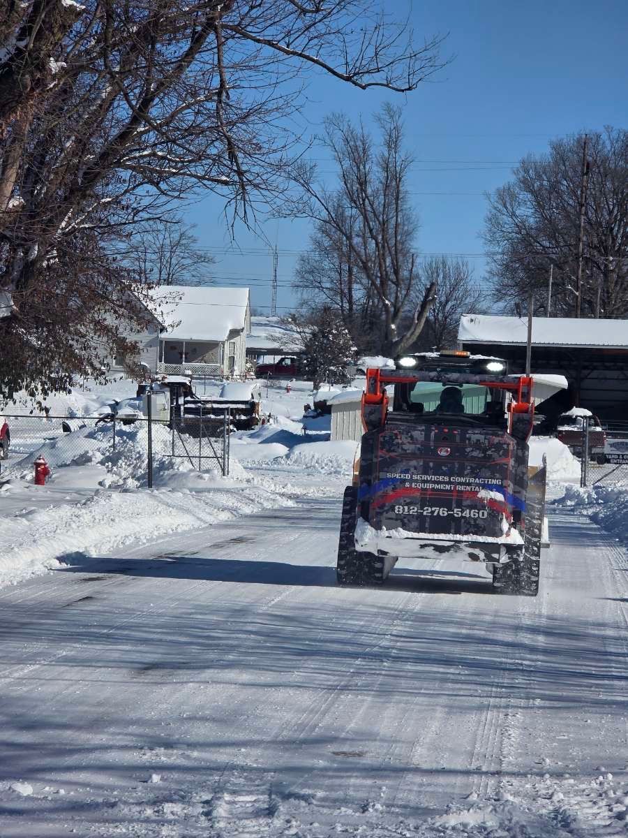 A skid steer clears snow from a residential street on a sunny winter day.